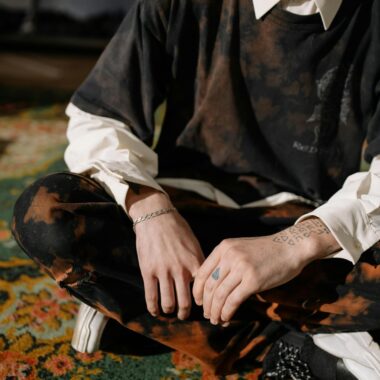 A stylishly dressed person with tattoos sitting indoors on a patterned carpet.